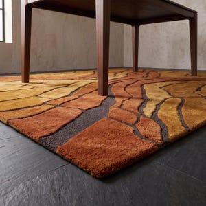 May include: A rectangular area rug featuring a textured, abstract pattern of organic, flowing shapes in shades of burnt orange, mustard yellow, and dark brown. The rug is positioned beneath the wooden legs of a table on a dark floor.