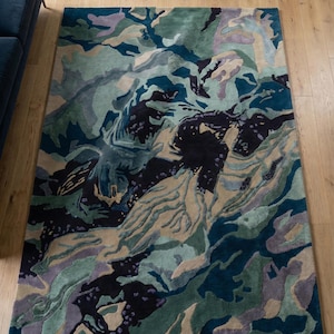 May include: A rectangular area rug with an abstract design. The rug features a blend of colors, including teal, green, beige, purple, and black, creating a textured, layered appearance. The rug is placed on a light-colored wooden floor.