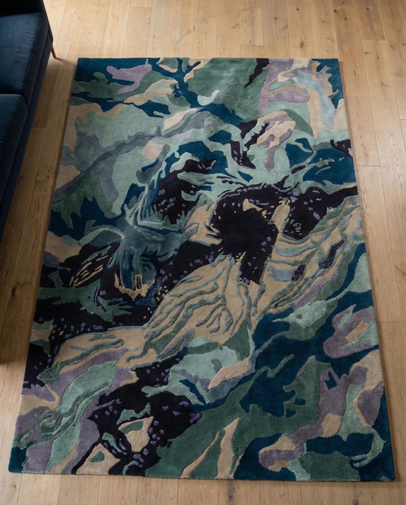 May include: A rectangular area rug with an abstract design. The rug features a blend of colors, including teal, green, beige, purple, and black, creating a textured, layered appearance. The rug is placed on a light-colored wooden floor.