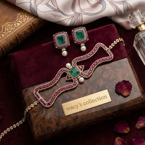 May include: A jewelry set featuring a necklace and matching stud earrings. The necklace has a geometric frame set with small red stones, a central emerald, and two white pearls. The earrings feature emeralds surrounded by red stones and pearls.