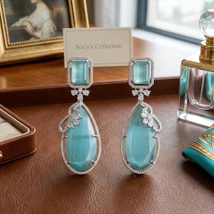 Puede incluir: Un par de elegantes pendientes colgantes con grandes piedras preciosas en forma de pera de color azul claro. Los pendientes tienen una piedra superior rectangular y están rodeados por un delicado engaste plateado con pequeñas piedras de acento transparentes.