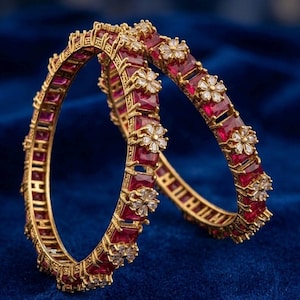 May include: Two gold-toned bangles featuring a series of rectangular, deep red stones set in a metal frame. Each bangle is accented with small, clear, flower-shaped clusters of stones spaced evenly along the outer edge of the circular band.