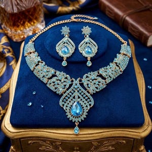 May include: A gold-coloured necklace and earring set adorned with light blue gemstones. The necklace has a floral design with a large teardrop pendant. The earrings have a matching design. The jewellery is displayed on a blue velvet surface.