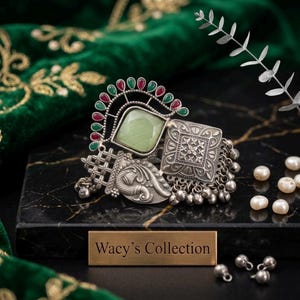 May include: An ornate silver ring with a large, light green square stone and smaller red and green stones. The ring has a detailed, textured design and is displayed on a black surface with the text "Wacy's Collection".