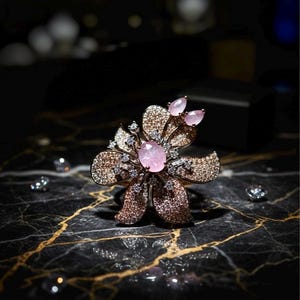 May include: A floral-shaped ring with a large oval pink gemstone center and smaller pink gems. The ring has a bronze-colored metal setting with sparkling accents. The ring is set on a black marble surface with gold veining.