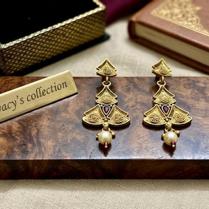 May include: Gold-toned earrings with a tree-like design, featuring red accents and a dangling pearl. The earrings rest on a wooden surface next to a gold nameplate that reads "wacy's collection". A decorative box and a book are in the background.