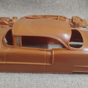May include: A tan plastic model car kit, featuring the body of a classic car. The kit includes the car's main body, roof, and other components, with some parts still in their original packaging. The car's design is reminiscent of vintage automobiles.