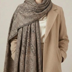 May include: A woman wearing a tan coat and a patterned scarf. The scarf features a detailed paisley design in shades of brown and black. The coat is a solid tan color, and the scarf is draped around the neck and shoulders.
