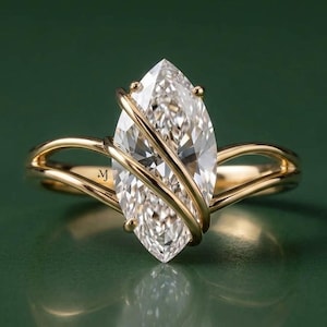 Puede incluir: Un anillo de oro con una gran piedra preciosa transparente de talla marquesa en el centro. La banda consta de dos hilos de oro curvos y delicados que envuelven la piedra, creando un diseño cruzado. Las letras MJ están grabadas en el interior del anillo.