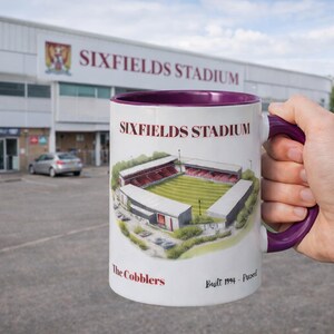 Peut inclure: Mug en céramique blanche avec intérieur et anse violets, représentant le stade de Sixfields. Le mug porte le nom du stade, ainsi que le texte "The Cobblers" et "Built 1994 - Present".