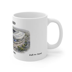 Pu&ograve; includere: Una tazza in ceramica bianca con una dettagliata illustrazione architettonica di uno stadio. Il testo riporta MADRID, SANTIAGO BERNABEU e Built 1947-Present. Il design mostra una vista aerea della struttura dello stadio con alberi.
