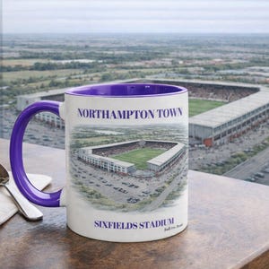 Peut inclure: Une tasse en céramique blanche avec une anse et un intérieur violets. La tasse présente une illustration du stade Sixfields et le texte "Northampton Town" et "Sixfields Stadium". Une cuillère et une serviette reposent sur une table en bois.