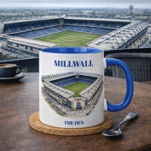 Pode incluir: Uma caneca de cerâmica branca com alça e interior azuis, com uma ilustração do estádio e os textos "MILLWALL" e "THE DEN". A caneca está sobre um descanso de copo de cortiça numa mesa de madeira.