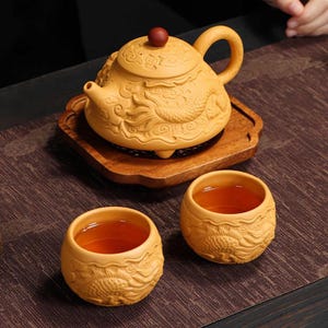 Op de afbeelding: Een gesneden houten theeset met een theepot met een drakenontwerp, een kleine houten dienblad en twee bijpassende theekopjes. De theepot heeft een rode knop op het deksel. De theekopjes zijn gevuld met thee. De set staat op een donkerbruin oppervlak.