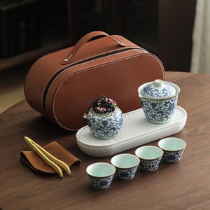 May include: A tea set with a brown leather case, a white tray, and blue and white porcelain items. The set includes a lidded tea cup, small cups, and a decorative jar with a floral pattern. The set is on a wooden surface.