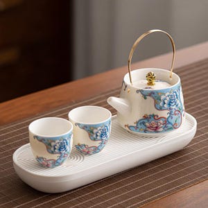 May include: A ceramic tea set with a teapot and two teacups, all decorated with blue and pink floral patterns on a cream background. The teapot has a gold handle and sits on a white tray.