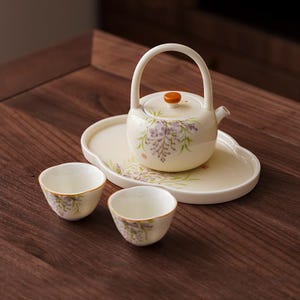 May include: A cream-colored ceramic tea set with a teapot, two cups, and a serving tray. The teapot has a curved handle and a lid with an orange knob. The set is decorated with purple floral patterns and green leaves.