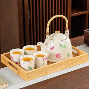 Peut inclure: Service à thé crème avec motifs floraux, comprenant une théière avec une poignée en bambou et quatre tasses assorties, présenté sur un plateau en bambou. Les tasses et la théière sont décorées de fleurs roses et de feuilles vertes.