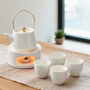 Op de afbeelding: Een witte keramische theepot met een goudkleurig metalen handvat en een gouden knop op het deksel, rustend op een bijpassende witte keramische theelichthouder met een brandend waxinelichtje, vergezeld door vier kleine witte keramische theekopjes.