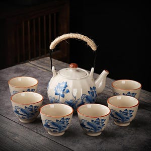 Puede incluir: Un juego de té de cerámica blanca con diseños florales azules. El juego incluye una tetera con asa de cuerda y seis tazas a juego. Las tazas y la tetera tienen un estampado floral azul y un borde rojo.