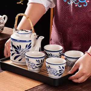 May include: A white ceramic tea set with blue floral patterns, comprising a teapot with a bamboo handle and four matching cups. The set is displayed on a black tray with a bamboo insert, ideal for serving tea.