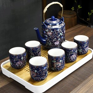 Puede incluir: Juego de té de cerámica azul con motivos florales. El juego incluye una tetera con asa y seis tazas a juego, todo dispuesto sobre una bandeja de bambú. Las tazas y la tetera tienen un borde dorado.