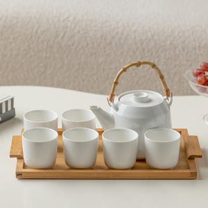 May include: A white ceramic tea set with a bamboo handle on the teapot. The set includes six matching cups and a bamboo serving tray. The tea set is ideal for serving tea or other hot beverages.