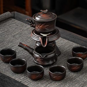 May include: A brown ceramic tea set with a teapot, a strainer, and six small cups. The teapot has a lid with a bird-shaped handle and bamboo leaf designs. The set is arranged on a gray surface.