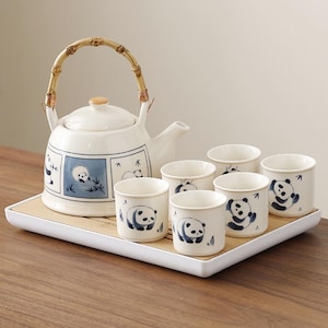 Puede incluir: Juego de té de cerámica blanca con ilustraciones de pandas. El juego incluye una tetera con asa de bambú y seis tazas a juego, todas decoradas con diseños de pandas azules. El juego se encuentra sobre una bandeja rectangular de bambú con borde blanco. La tetera tiene un pomo de madera.