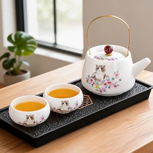 May include: A white ceramic tea set with a teapot and two teacups, decorated with a cat and floral pattern. The teapot has a gold handle and a red lid knob. The set is on a black tray, and the teacups are filled with tea.