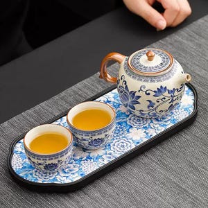 May include: A traditional tea set with a teapot and two teacups filled with tea, on a floral patterned tray. The teapot and cups have a blue and white floral design. The tray is approximately 30 cm long.
