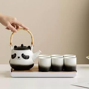 Könnte beinhalten: Panda-Teeservice mit einer weiß-schwarzen Teekanne mit Bambusgriff und sechs passenden Tassen. Das Set wird auf einem weißen Tablett mit Bambuseinsatz präsentiert, ideal zum Servieren von Tee.