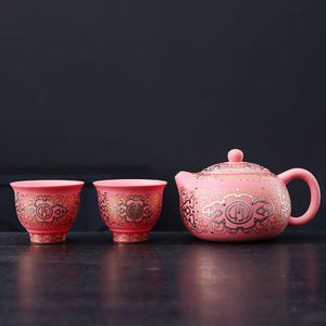 Może przedstawiać: Różowy ceramiczny zestaw do herbaty ze złotymi wzorami dekoracyjnymi. Zestaw zawiera czajniczek z pokrywką i dwie pasujące filiżanki. Zestaw do herbaty jest prezentowany na ciemnej powierzchni.