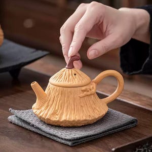 Könnte beinhalten: Eine hellbraune Keramik-Teekanne mit einem strukturierten, strohähnlichen Design. Die Teekanne hat einen gebogenen Griff, einen Ausguss und einen Deckel mit einem kleinen, braunen Knopf. Sie steht auf einem grauen Tuch.