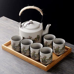 May include: A light grey ceramic tea set comprising a teapot with a woven handle and six cups. Each piece features a landscape design. The set is presented on a rectangular wooden tray.