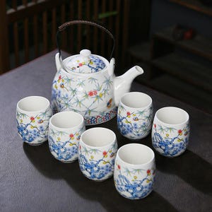 Może przedstawiać: Biały ceramiczny zestaw do herbaty z czajnikiem i sześcioma pasującymi filiżankami. Czajnik ma czarny uchwyt i pokrywkę. Zestaw jest zdobiony niebieskimi wzorami kwiatowymi i bambusowymi, z czerwonymi i żółtymi akcentami. Filiżanki są cylindryczne.