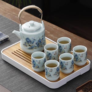 Puede incluir: Juego de té de cerámica con una tetera y seis tazas a juego, decoradas con un diseño de paisaje montañoso azul y blanco. La tetera tiene un asa de cuerda. El juego se presenta en una bandeja de bambú.