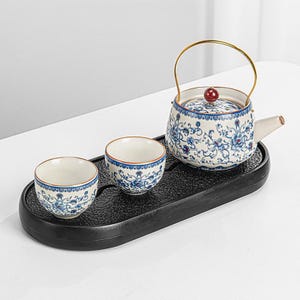 Może przedstawiać: Ceramiczny zestaw do herbaty z imbrykiem i dwoma filiżankami, zdobiony niebieskimi wzorami kwiatowymi na białym tle. Imbryk ma złoty uchwyt i czerwony guziczek na pokrywce. Zestaw stoi na czarnej tacy.
