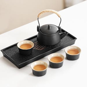 May include: A black ceramic tea set with a teapot, three teacups, and a serving tray. The teapot has a woven handle. The teacups and teapot are filled with tea. The tray has a textured surface.