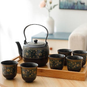 May include: A black ceramic tea set with a teapot and six matching cups, all decorated with gold floral patterns. The teapot has a curved handle and a lid. The set is displayed on a wooden tray, perfect for serving tea.