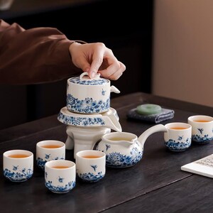 Puede incluir: Un juego de té de porcelana blanca y azul con un diseño floral. El juego incluye una tetera, tazas y un infusor de té. Las tazas de té están llenas de té. El juego de té está sobre una mesa de madera oscura.