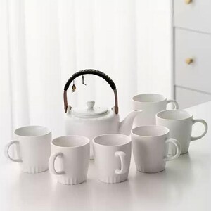 Puede incluir: Una tetera de cerámica blanca con un asa negra y varias tazas blancas. La tetera tiene un asa decorativa y una tapa. Las tazas son sencillas y cilíndricas, con un asa en el lateral. El conjunto está dispuesto sobre una superficie blanca.