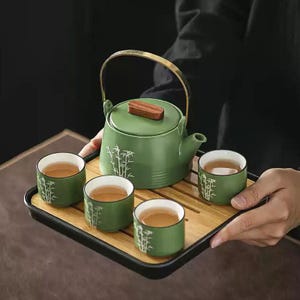 Op de afbeelding: Een groene keramische theeservies met een theepot en vier kopjes, elk versierd met een bamboe-ontwerp. De theepot heeft een houten deksel en een gouden handvat. De set wordt gepresenteerd op een bamboe dienblad.