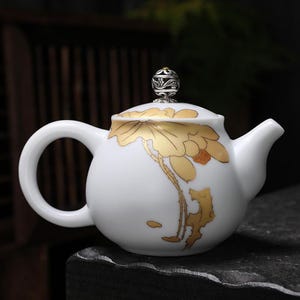 May include: White ceramic teapot with a gold lotus flower design. The teapot has a rounded body, a curved handle, and a spout. The lid is topped with a decorative silver and gold knob. The teapot is likely used for serving tea.