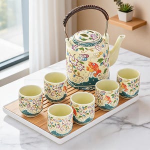 Op de afbeelding: Een keramische theeset bestaande uit een theepot met een gevlochten handvat en zes bijpassende kopjes, versierd met kleurrijke koikarpers, bloemmotieven en gestileerde wolken op een crèmekleurige achtergrond, op een rechthoekig bamboe dienblad.