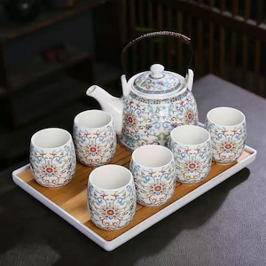 Könnte beinhalten: Ein weißes Keramik-Teeservice mit einem Blumenmuster in Rot, Blau und Gelb. Das Set enthält eine Teekanne mit Henkel und Deckel sowie sechs passende Teetassen. Das Set wird auf einem rechteckigen Bambustablett präsentiert.