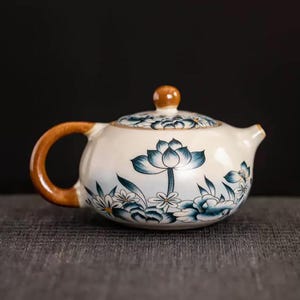 May include: A white ceramic teapot with a brown handle and lid knob. The teapot is decorated with blue floral designs, including lotus flowers and other blossoms. The spout and handle are brown.
