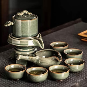 May include: A celadon green tea set with a crackle glaze. The set includes a tea pot with a lid, a serving pitcher, and six small tea cups. The tea cups are filled with tea. The tea set is on a dark surface.