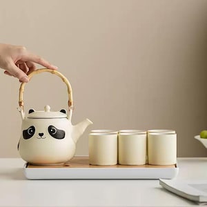 Könnte beinhalten: Eine cremefarbene Teekanne im Panda-Design mit Bambusgriff, zusammen mit vier passenden Teetassen. Die Teekanne hat ein Panda-Gesicht mit schwarzen Augen und Ohren. Das Set wird auf einem rechteckigen Tablett präsentiert.