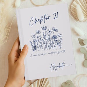 May include: A white book cover with the text "Chapter 21" in navy blue script, along with a floral illustration and the words "A new chapter, endless growth." The name "Elizabeth" is signed below the text.